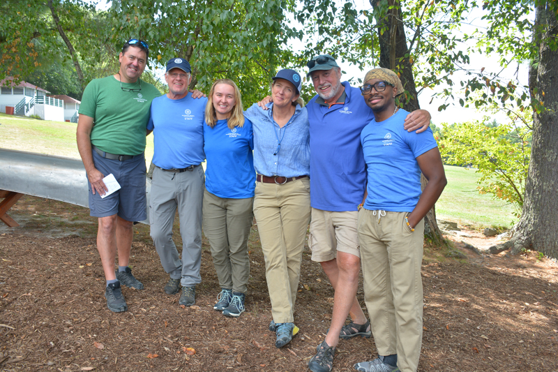 The image shows a group of six people posing for a photo outdoors. They are all smiling and appear to be in a relaxed setting, possibly a park or recreational area. The people are dressed in casual attire, with some wearing matching shirts. The background includes trees and a glimpse of a building.